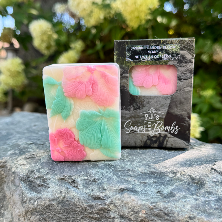 A rectangular bar of soap with embossed pink and mint green hibiscus flowers on a cream base, standing next to its black branded retail box outdoors on a stone surface with blurred green foliage in the background.
