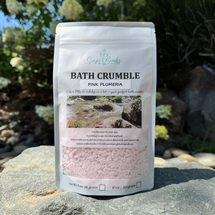 Bath Crumble packaging labeled 'Pink Plumeria' by Soaps & Bombs on a rock with plants in the background