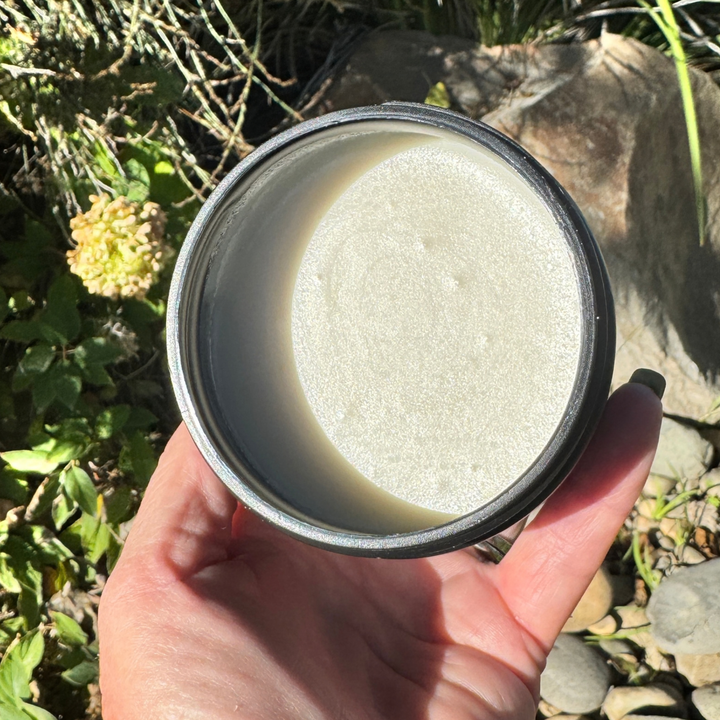 A hand holding the open lid of a black metal container outdoors, showing a pale off-white cream or balm-like substance inside. The background features green foliage, rocks, and natural sunlight.